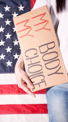 Young woman protester holds cardboard with My Body My Choice sign against USA flag on background....