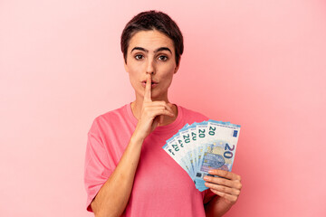 Young caucasian woman holding banknotes isolated on pink background keeping a secret or asking for silence.