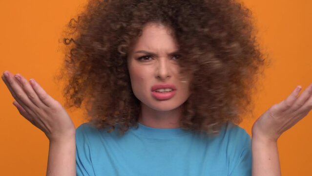 Displeased curly haired blonde woman wearing blue t-shirt touching her hairstyle in the orange studio