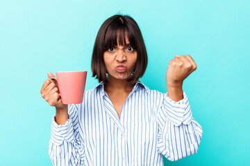 Young mixed race woman holding a pink mug isolated on blue background showing fist to camera,...