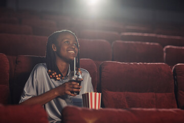 African happy woman with popcorn and soda enjoying watching of movie in the cinema
