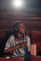 African young woman watching comedy in the cinema, she sitting on armchair with popcorn and soda and smiling