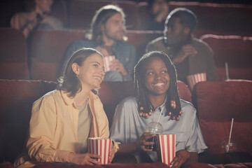 Two happy women eating popcorn and watching comedy on comfortable chairs in auditorium