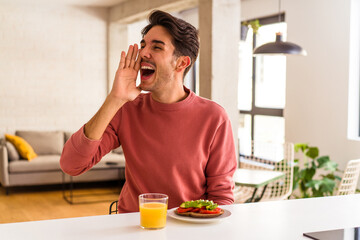 Fototapeta na wymiar Young mixed race man having breakfast in his kitchen shouting and holding palm near opened mouth.