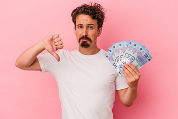 Young caucasian man holding banknotes isolated on pink background showing a dislike gesture, thumbs...
