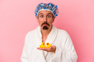 Young caucasian man going to the shower with rubber ducks isolated on pink background shrugs...