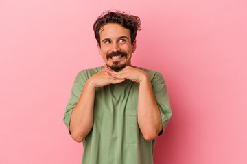 Young caucasian man isolated on pink background keeps hands under chin, is looking happily aside.