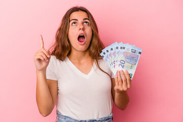 Young caucasian woman holding bills isolated on pink background  pointing upside with opened mouth.