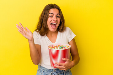 Young caucasian woman holding a popcorns isolated on yellow background  receiving a pleasant surprise, excited and raising hands.