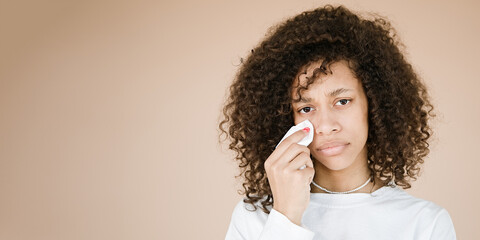 Upset doleful young african american woman sobs and cries from despair wipes tears away with a tissue isolated over beige background with copy space for your advertising content.           