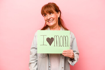 Middle age caucasian woman holding I love mom placard isolated on pink background looks aside smiling, cheerful and pleasant.