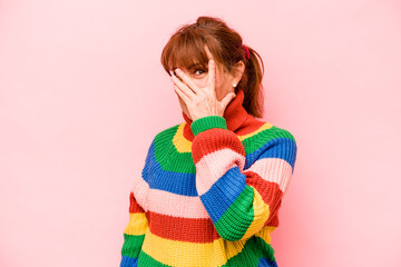 Middle age caucasian woman isolated on pink background blink at the camera through fingers, embarrassed covering face.