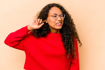 Young hispanic woman isolated on beige background covering ears with fingers, stressed and desperate by a loudly ambient.