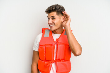 Young caucasian man wearing life jacket isolated on white background trying to listening a gossip.