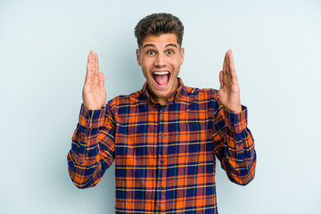 Naklejka premium Young caucasian man isolated on blue background receiving a pleasant surprise, excited and raising hands.