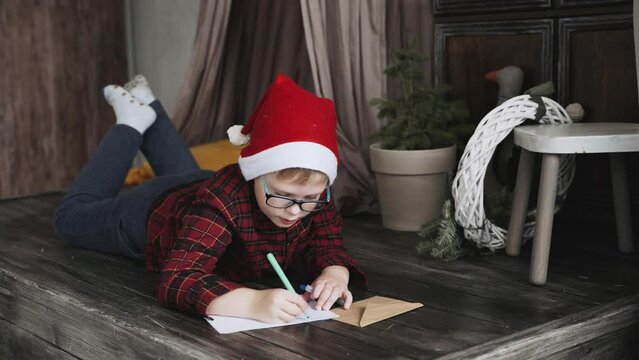 Cute boy in a Christmas hat and glasses lies on the wooden floor and diligently writes a letter to Santa Claus with felt-tip pen. Belief in miracles, Christmas miracle, cherished wishes.