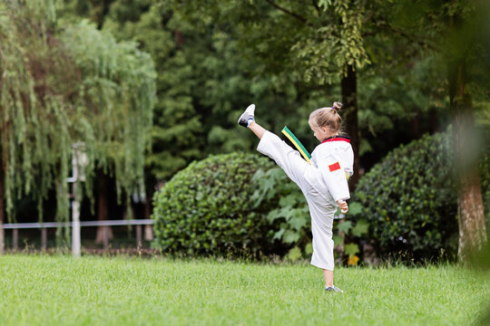 Caucasian little girl seven years old in kimono with yellow green belt exercising Taekwondo at summer park alone during coronavirus covid-19 lockdown, self isolation and social distancing