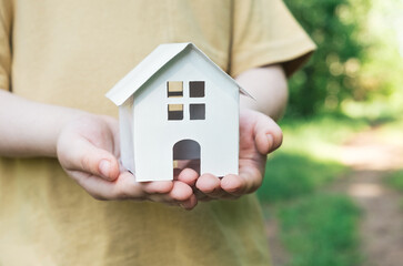 Child holding small house model on green forest background. Eco house concept, family life on nature, sustainability. Closeup