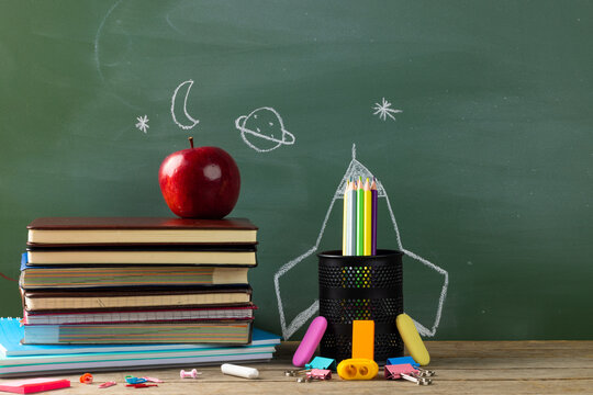 Image of school supplies, apple and notebook over drawings on black board