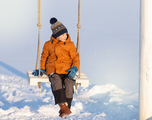 boy sits on a swing in winter and looks into the snowy winter distance
