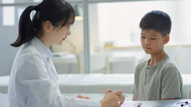 Female doctor checking neck of Asian tween boy, telling diagnosis and taking notes during consultation in clinic