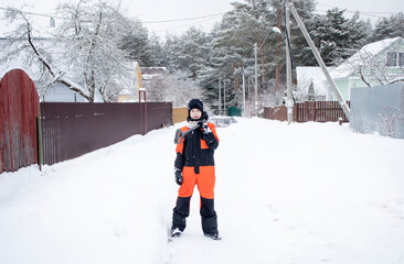 a boy in an orange jumpsuit stands on a village street with a large snow shovel in winter