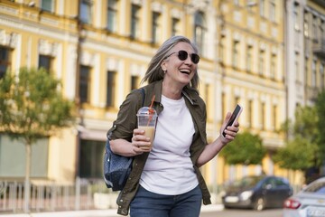 European senior woman with gray hair happy enjoying free time after work or traveling holding phone while having juice on the go using plastic cup in city background. Enjoying life mature woman