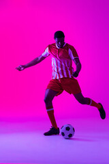 African american male soccer player kicking football over neon pink lighting