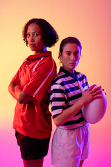 Portrait of two diverse female rugby players with rugby ball over neon pink lighting