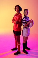 Portrait of two diverse female rugby players with rugby ball over neon pink lighting