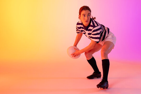 Caucasian female rugby player crouching with rugby ball over neon pink lighting - Powered by Adobe