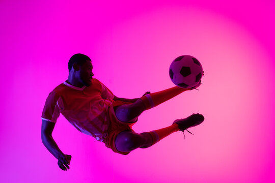 African american male soccer player kicking football over neon pink lighting