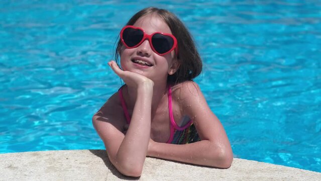 Happy cute little girl in heart shaped sunglasses in swimming pool on sunny day. Childhood, valentine's day and summer vacation concept. Smiling child having fun outdoors on family holidays