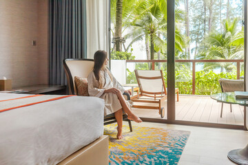 Woman on vacation in tropical luxury resort. Female traveler in bathrobe sitting on chair in hotel room enjoy palm trees and sea view. Summer holidays in modern hotel. - obrazy, fototapety, plakaty