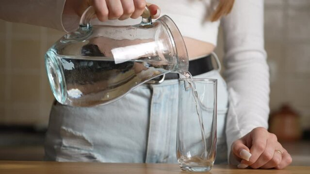 Female hands pouring water into glass from jug at kitchen background, close up. Unrecognizable person drinking water at home kitchen. Unknown girl standing in home alone