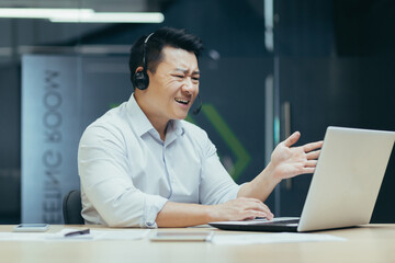 Angry and annoyed asian businessman with headset for video call, talking in online meeting, looking at laptop screen, man at work