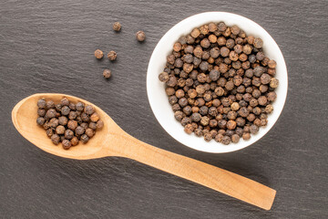 Dry fragrant black pepper in a white saucer with a wooden spoon on a slate stone, close-up, top view.