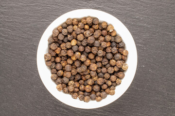 Dry fragrant black pepper in a white saucer  on a slate stone, close-up, top view.