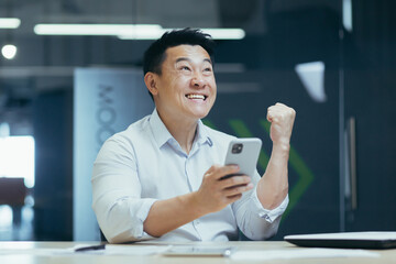 Happy and successful Asian businessman working in office, typing on phone and reading good news, celebrating triumph and victory