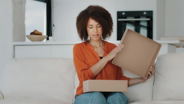 Curious African American buyer customer woman with curly hair sitting at couch at home unbox opening unpacking parcel box gift present purchase order from online shop shipping delivery service indoors