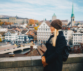 Young beautiful pretty blonde tourist girl in warm hat and coat with backpack walking at cold autumn in Europe city enjoying her travel in Zurich Switzerland