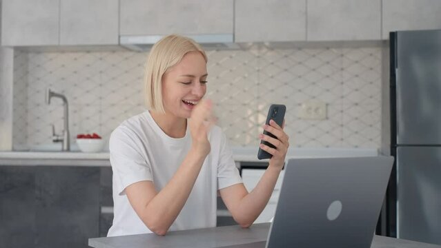 Joyful blonde woman waves bye in video call when sitting at laptop in kitchen