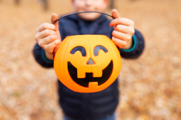 Child holds a bucket shaped like a halloween pumpkin jack o lantern in autumn park. Kid with...