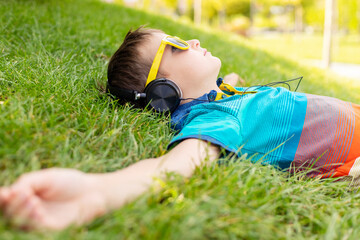Happy boy in sunglasses lying on the grass in the park and listening to music in headphones on a...