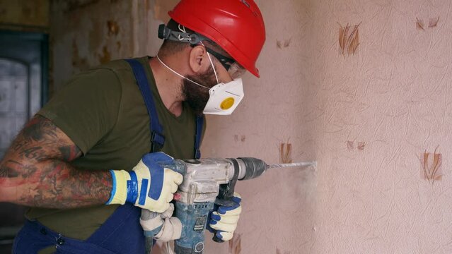 Builder working with rotary hammer drill. Contractor drills holes in concrete with jackhammer to demolish the wall for apartment remodelling and renovation. Electrician in uniform installs wiring.