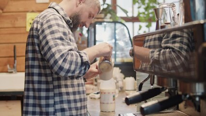Mid shot of professional male bearded barista wearing checkered shirt pouring warm milk in cup with coffee to make cappuccino, working in a cafe, preparing coffee with coffee machine at counter coffee