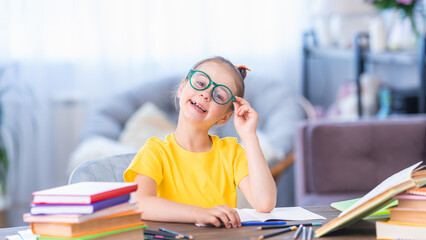 Cute happy schoolgirl with glasses. He sits at his desk at home and writes in a notebook. Back to school. The little girl is happy to get knowledge and do her homework. Family education
