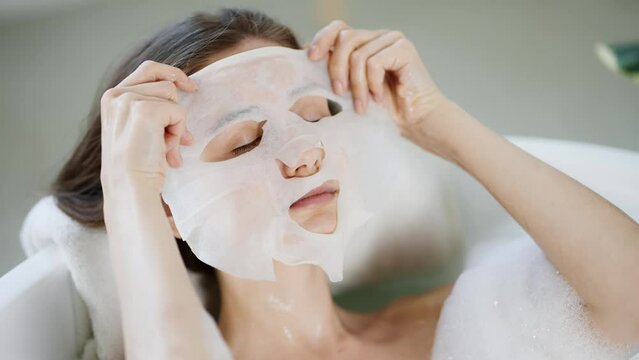 Young domestic woman posing apply tissue face mask for skin care while laying in the bath