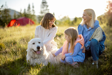 Portrait of two women with little girl and white dog sit together on nature during sunset. Homosexual relationship, parenting kid and travel in mountains concept
