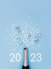 Open bottle of champagne on light blue background decorated with silver star confetti and numbers 2023. New Year celebration or party. Selective focus.